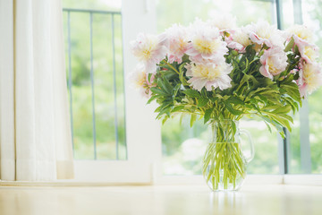 Big beautiful pale pink peonies bouquet in glass vase over window background. Light Home  decoration with flowers and vase. Living room interior