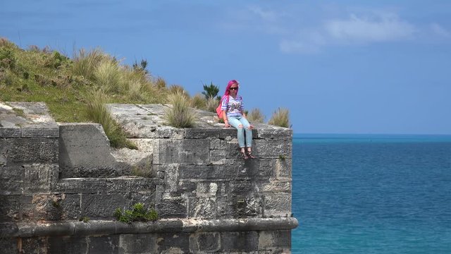Girl with pink hairs sits on the edge of the fort wall and looks at the sea. Royal Naval Dockyard, Bermuda