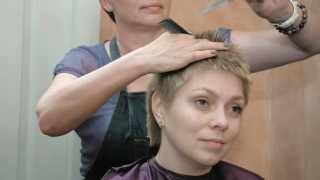 Hairdresser cuts combs and styles pretty blond woman short hair with scissors comb and various tools. Beauty spa and makeup salon studio scene. 4K UHD video footage.