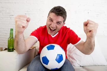 Foto auf Acrylglas man watching football on tv wearing team jersey celebrating goal happy on sofa © Wordley Calvo Stock