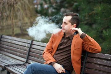 A young man sits on a bench and smokes an electronic cigarette.