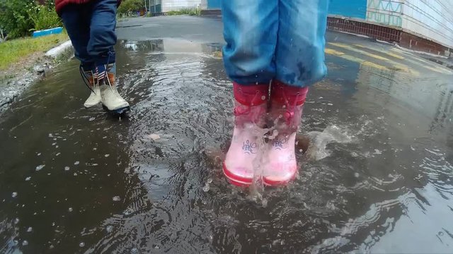 Child in boots walking and jumping in muddy puddle after rain.
