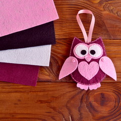 Funny maroon felt owl with a pink heart. Gift idea for Valentine's day, 8 March. Cheap and easy decorating idea. Sewing workplace. Burgundy, pink, white and black felt texture. Brown wooden background
