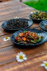 Different kinds of tea on black plates on wooden table with flowers of chamomile in garden and on nature background. Assortment of dry tea. Tea concept. Tea leaves. Closeup