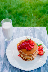 Pancakes with fresh strawberry and jem near glass with milk on white plate on white wooden background  in garden or on nature background.  Stack of pancakes on the table.