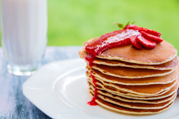 Pancakes with fresh strawberry and jem near glass with milk on white plate on white wooden background  in garden or on nature background.  Stack of pancakes on the table.
