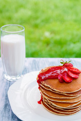 Pancakes with fresh strawberry and jem near glass with milk on white plate on white wooden background  in garden or on nature background.  Stack of pancakes on the table.