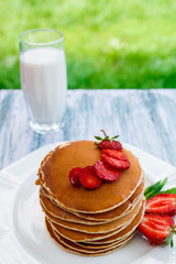 Pancakes with fresh strawberry and mint near glass with milk on white plate on pink wooden background  in garden or on nature background.  Stack of pancakes on the table.