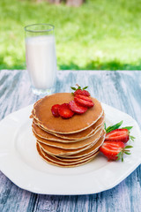 Pancakes with fresh strawberry and mint near glass with milk on white plate on pink wooden background  in garden or on nature background.  Stack of pancakes on the table.