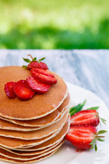 Pancakes with fresh strawberry and mint on white plate on pink wooden background  in garden or on nature background.  Stack of pancakes on table.