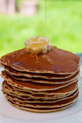 Pancakes with butter and honey on white plate on  in garden or on nature background. Maple syrup pouring on pancakes stack.