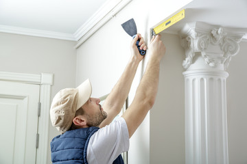 Worker holds putty knife and measures the wall corner using metal angle.  Finishing work.