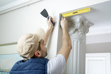 Worker holds putty knife and measures the wall corner using metal angle.  Finishing work.
