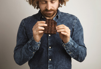 Young happy bearded man with healthy skin and curly hair dreamely holds organic freshly baked chocolate bar with both hands carefully with closed eyes