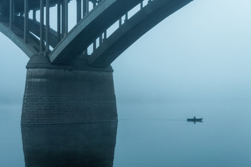 fisherman on a small boat sails next to the pillars of the bridge in the fog.