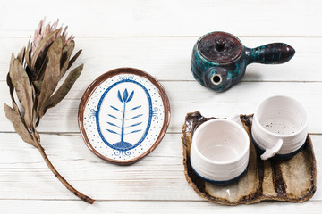Close-up of clay tea set composition. Teapot, two cups on saucer, plate and rare flower served on white wooden table. Clay tea set for two in village style .