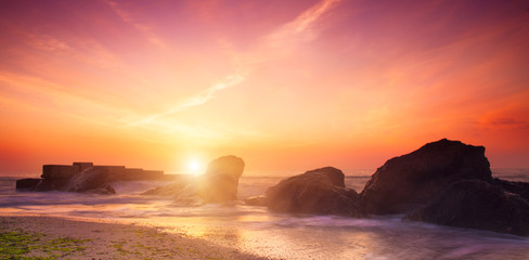 Fototapeta na wymiar Summer seasonal natural vacation background. Romantic morning at sea. Big boulders sticking out from smooth wavy sea. Pink horizon with first hot sun rays. Long exposure. 
