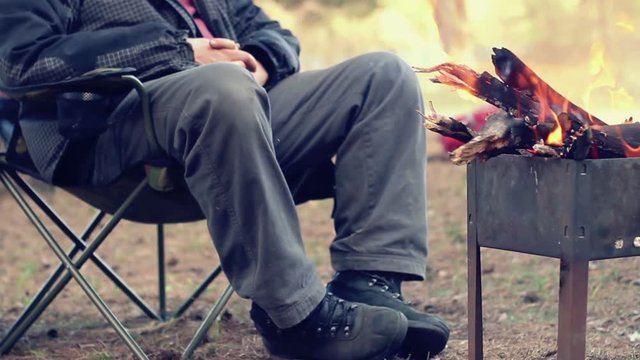 Tourist sitting on camp chair near campfire. Outdoor recreation. Bonfire in forest. Man resting near fire. Bonfire in camp. Rest in camping. Relaxing in forest. Relax near wood burning in mangal