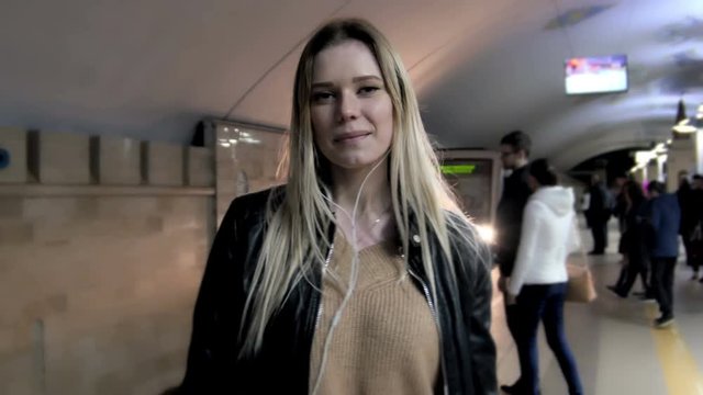 Сute girl with long blonde hair in leather jacket with straightens hair standing in metro against the background of a train coming