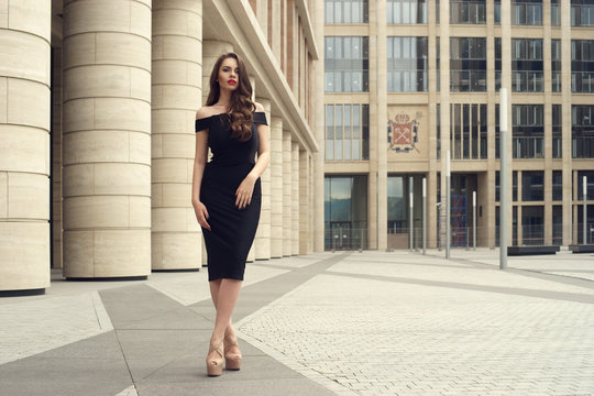 Young elegant girl posing at city street. Pretty beautiful business woman in elegant black dress against city background. Full length horizontal portrait.