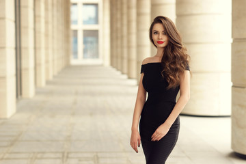 Young elegant girl posing at city street. Pretty beautiful business woman in elegant black dress against city background.