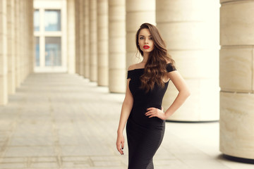 Young elegant girl posing at city street. Pretty beautiful business woman in elegant black dress...