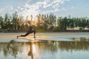 Woman practicing Warrior yoga pose outdoors over sunset sky back