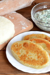Fried pies on a plate, glass bowl of curd, a spoon, rolling pin, dough, cutting board. Simple appetizer recipe. Easy vegetarian snack idea. Homemade pastry. Vertical photo. Rustic style. Macro