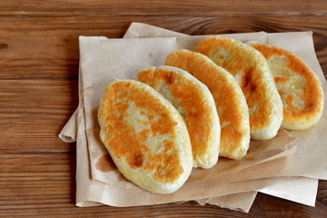 Five pies on paper and on wooden table. Fried pies with cottage cheese filling. Patties cooked on a yeast-free dough. Delicious hearty snack, breakfast, lunch. Homemade fast food. Frugal recipe idea