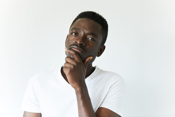 Handsome African man in white T-shirt, looking at the camera with thoughtful skeptical expression, holding hand on his chin. Black male with mistrustful look posing against white concrete wall