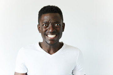 Headshot of annoyed stressed African American man in casual clothes, looking at the camera with irritated and angry expression, clenching his ultrawhite teeth in exasperation against white studio wall