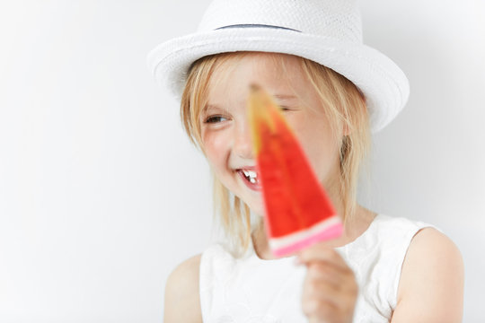 Smiling Caucasian Child Laughing And Hiding Behind Red Popsicle In Her White Beachwear Summer Clothes. Small Blond Girl Looking Sideways In Playful Manner Of Happy-go-lucky Kid.