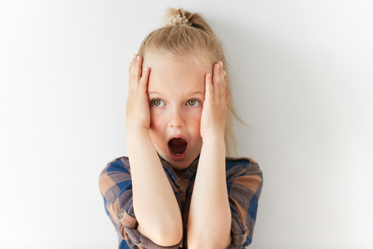 Blond Caucasian Female Child Opening Her Mouth In Astonishment And Clutching Head In Horror. Scared And Terrified Kid Is Frightened By Negative News And Ready To Scream.