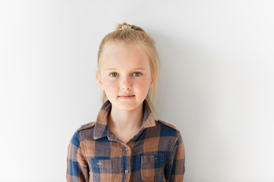 Cute blond girl with posing on white background. Portrait of relaxed kid with nice kind look. Calm and pretty little Caucasian girl with pony-tail in brown shirt. Childish innocence