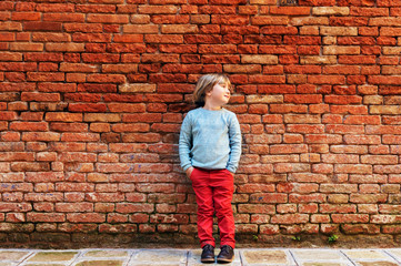 Little stylish blond boy posing against red brick wall, wearing terracotta trousers, blue pullover and brown vintage shoes