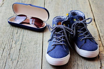Fashion background. Denim sneakers for a boy and sunglasses in a case on a wooden background