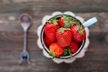 Fresh juicy strawberries in old rusty mug. Rustic wooden background with hand made crochet napkin and vintage spoon. Top view, place for text.