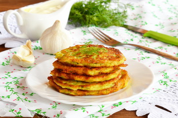 Pancake made from zucchini, eggs, flour, salt, garlic, dill. Delicious zucchini fritters on a plate. Fork, knife, green dill, gravy boat, garlic, napkin. Easy and healthy zucchini recipe.