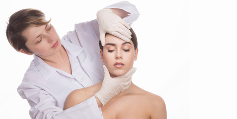 Examining face concept. Portrait of young beautiful woman sitting with her eyes closed while professional cosmetologist examining her face over white background.