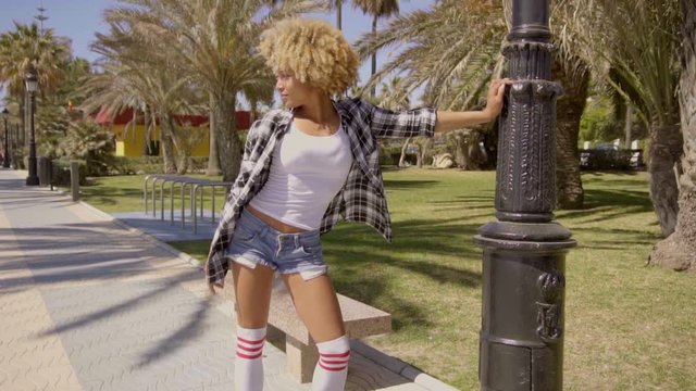 Wide shot of young black lady dressed in plaid men's shirt and sexy white top while waiting in the park for somebody and looking around in slow motion.