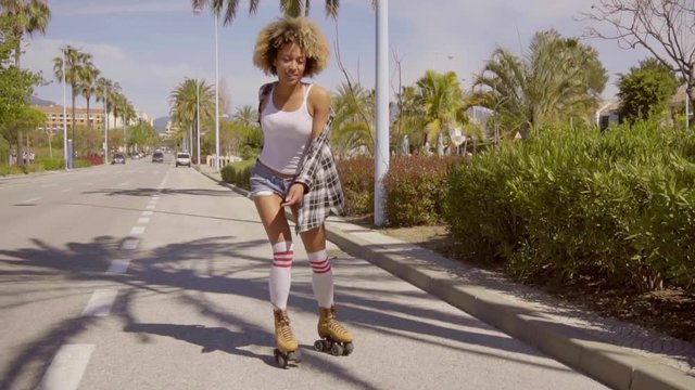 Young black woman wearing blue jean shorts roller skating along the road with palm trees in the distance