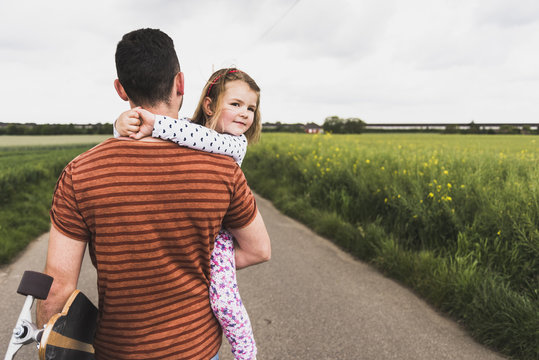 Father with skateboard carrying daughter on country lane