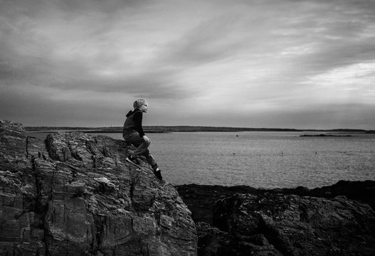 Young boy sitting on large rock beside lake, looking at view, black and white