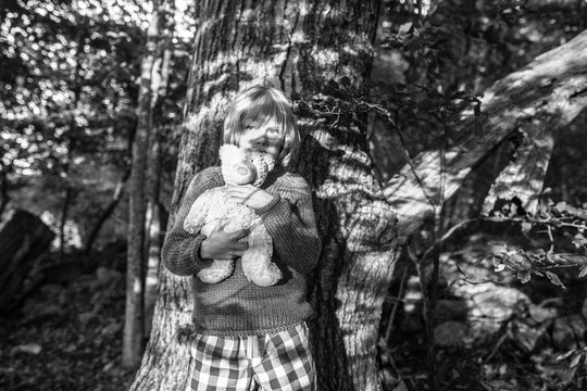 Young boy leaning against tree, holding soft toy, black and white