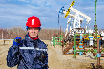 Portrait of man engineer in the oil field wearing red helmet and work clothes holding wrenches in his hand and radio in jacket pocket. Blurry pump jack and wellhead background. Oil and gas concept. 