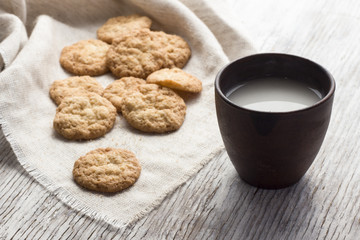Oatmeal cookies on a linen a napkin with milk in ceramic cup on a light wooden background. Breakfast in the village.