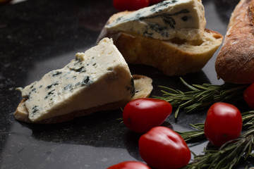 Blue cheese with french baguette, tomato and herbs on black marble table. Traditional snacks in France and Italy.
