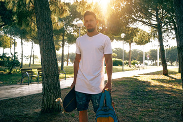 Young guy in a blank white t-shirt and shorts is standing in the park on a plant background. A man with a skateboard and a backpack is looking at camera. Flare light.