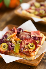 Hausgemachtes frischgebackenes Pizzastück belegt mit Schinken, Salami, Oliven, Paprika und Käse auf Holzteller serviert (Selektiver Fokus, Fokus auf die grüne Olive rechts)