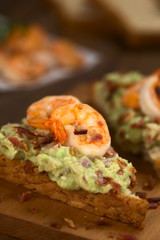 Vollkorntoastbrot belegt mit Guacamole (Avokado-Creme), gebratenen Garnelen und Speckstückchen (Selektiver Fokus, Fokus auf die Vorderseite der Garnele auf dem Brot)